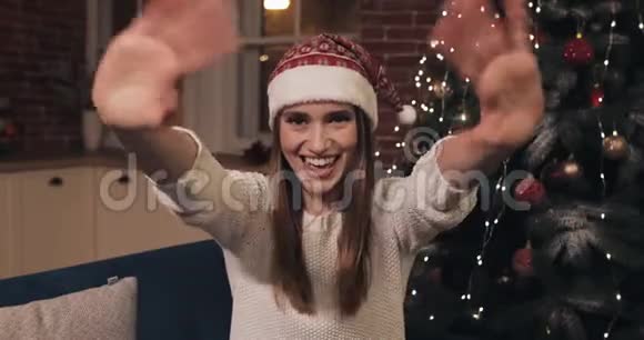 幸福的女人戴着桑塔的帽子坐在家中的圣诞树附近挥舞着嗨拿着礼物盒摇着礼物盒问候问候视频的预览图