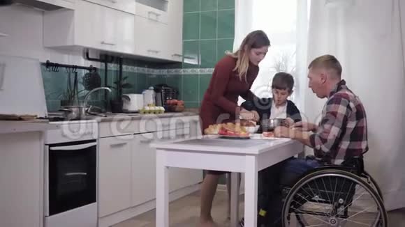 友好的家庭男父亲坐在轮椅上坐在厨房的餐桌上和他的儿子和妻子一起准备晚餐说话很友好视频的预览图