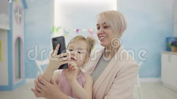 年轻的妈妈学习她的小女儿在智能手机上自拍视频的预览图