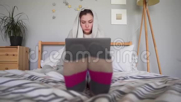 一个穿着灰色袜子的年轻女人在一台笔记本电脑上工作她裹着个格子里坐在家里的床上视频的预览图