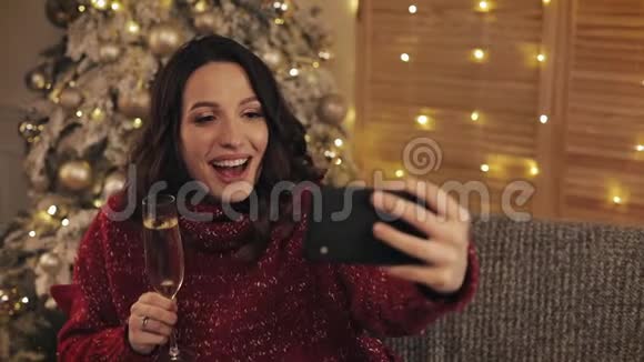 年轻的微笑美丽的黑发女人拿着香槟酒杯坐在圣诞树旁在圣诞树旁闪耀着灯光视频的预览图