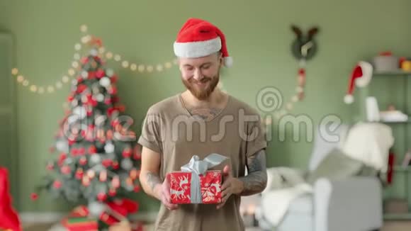 年轻英俊的留着胡须的纹身男穿着绿色t恤戴着红色圣诞帽正在家里给相机送礼物视频的预览图