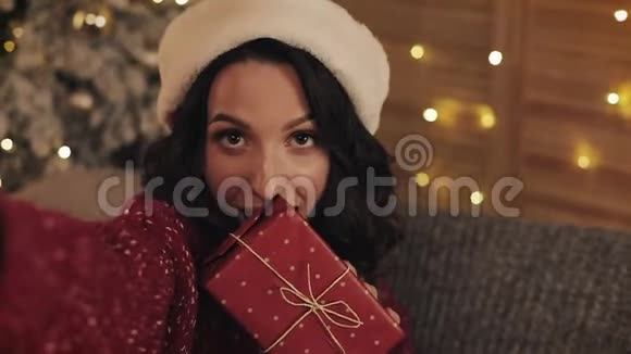 可爱的年轻的布鲁内特女士带着圣诞帽坐在沙发上靠近圣诞树在舒适的家背景下举行视频的预览图