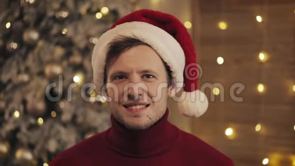 室内特写照片英俊的微笑男子戴着桑塔斯帽子坐在圣诞树附近的沙发上闪闪发光视频的预览图