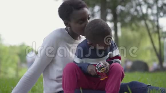 小可爱的非裔美国男孩在公园里玩塑料玩具坐在妈妈圈上女人和视频的预览图