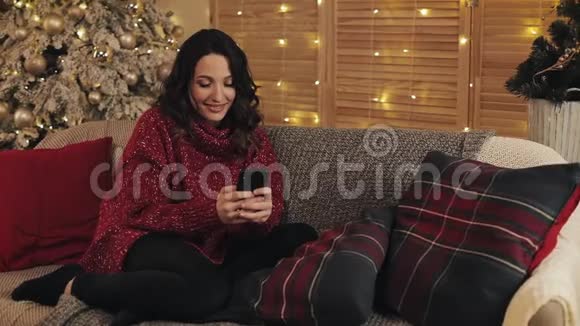 穿红汗衫的年轻美丽的笑脸女人用她的智能手机打字聊天坐在圣诞树附近视频的预览图