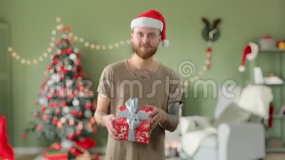年轻英俊的留着胡须的纹身男身穿绿色t恤戴着红色圣诞帽正在家带着礼物跳舞视频的预览图