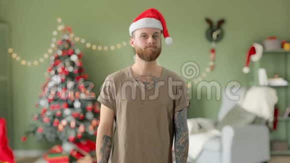 年轻英俊的长胡子纹身男身穿绿色t恤戴着红色圣诞帽正带着圣诞礼物回家视频的预览图