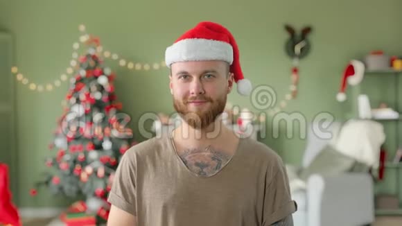 年轻英俊的留着胡须的纹身男身穿绿色t恤戴着红色圣诞帽家里还有圣诞装饰品视频的预览图