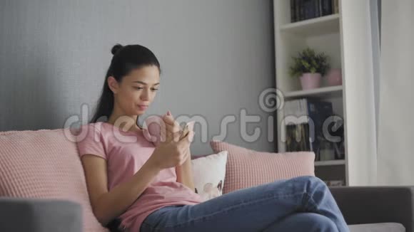 美丽的年轻女性使用智能手机在家里的沙发上放松上网浏览短信喝咖啡享受享受视频的预览图