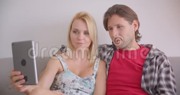 一对有魅力的白种人成年夫妇在平板电脑上打电话坐在沙发上挥手致意视频的预览图