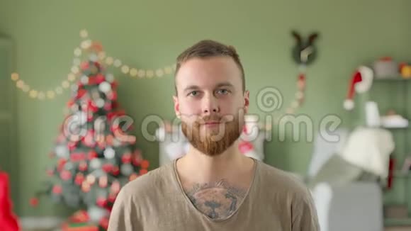 身穿绿色t恤长胡子纹身的年轻帅哥戴着一顶红色的圣诞帽带着圣诞节在家微笑视频的预览图