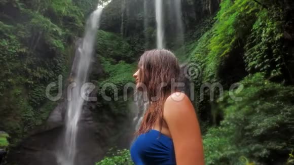 一个在瀑布背景下的热带雨林中的年轻女孩站在雨中与自然的统一视频的预览图