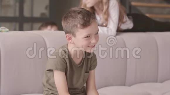 笑的小男孩坐在前景的沙发上的肖像姐姐和哥哥在后台玩视频的预览图