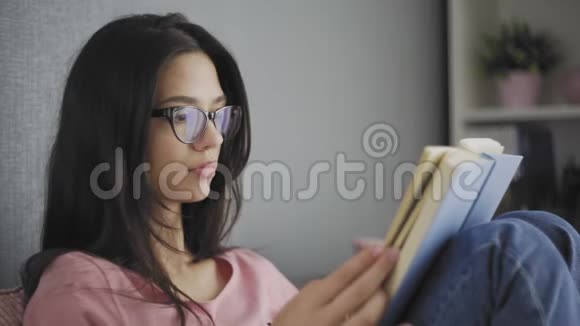 漂亮快乐的女学生戴着眼镜在一所现代化的房子的沙发上看书视频的预览图