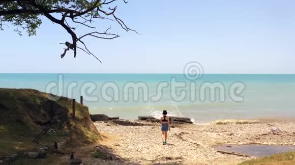 穿着蓝色短裤的女孩在沙滩上走向大海海浪拍打海岸视频的预览图