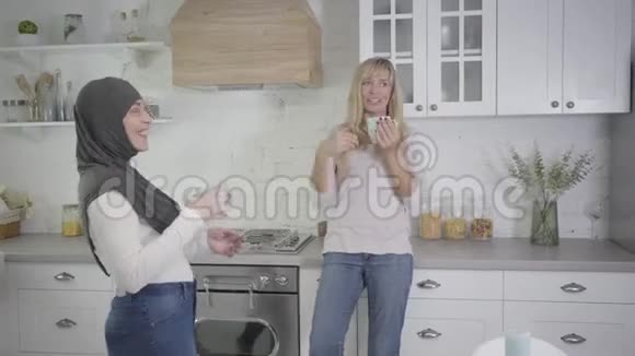 金发白种人女人和她的女性穆斯林朋友在现代厨房喝茶或咖啡笑朋友视频的预览图