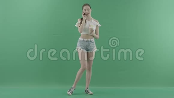 年轻健康的女性在绿色背景下喝鸡尾酒时会通过智能手机打电话视频的预览图