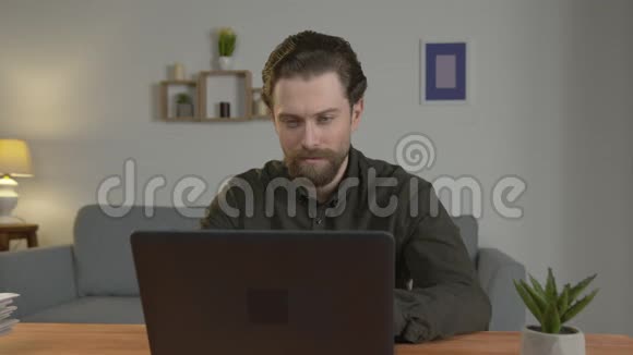 一个留着胡子的白人坐在桌子旁在笔记本电脑上工作在家工作看着相机严肃视频的预览图