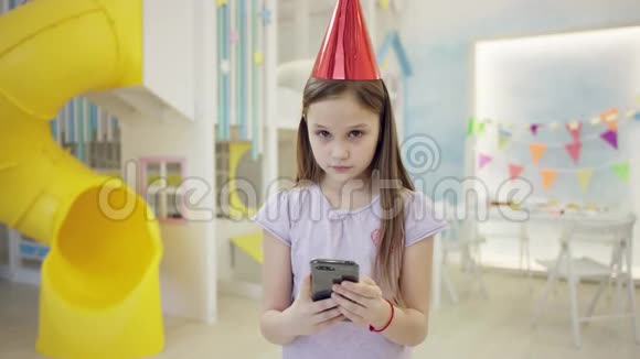 戴着生日帽的小女孩用智能手机在镜头前抚摸头发视频的预览图