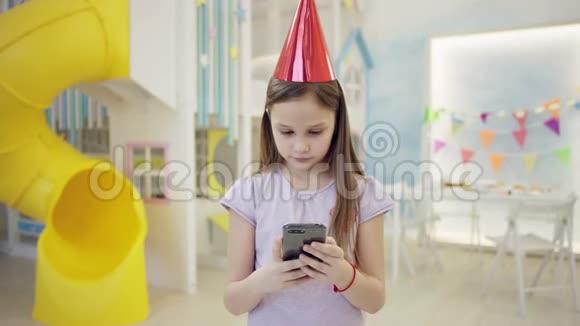 戴着生日锥帽的漂亮小女孩在镜头前的智能手机上发短信视频的预览图