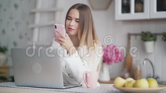 年轻女子坐在餐桌上厨房里拿着笔记本电脑用手机上网聊天视频的预览图