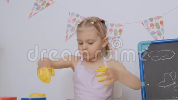 一个小可爱的女孩坐在孩子们的桌子旁用手指画画把她的手染成黄色视频的预览图