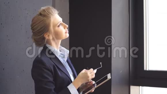 年轻的商务女士站在一间现代化的办公室的窗口用平板电脑做笔记视频的预览图