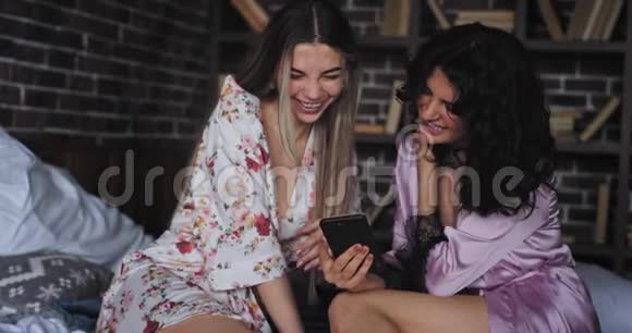 两个穿着睡衣的有魅力的大女人笑着一起看着智能手机兴奋又兴奋视频的预览图