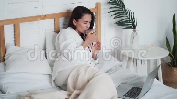 穿着白色浴袍的女人躺在床上喝茶在笔记本电脑上看电影视频的预览图
