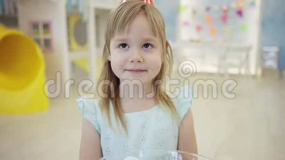 漂亮的小女孩正在游戏室里吃一个棉花糖头上戴着一顶锥帽在摄像机前视频的预览图