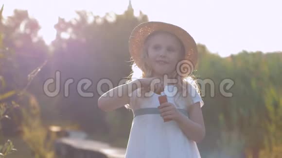 戴着草帽的快乐美丽微笑的小女孩在阳光下吹泡泡和欢笑视频的预览图