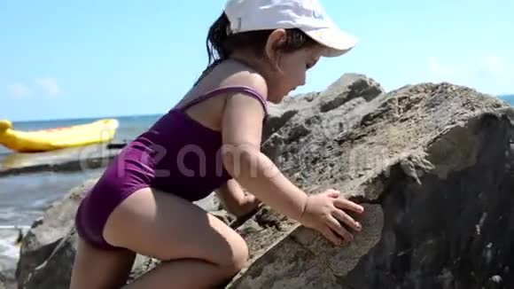 穿着紫色泳衣的小可爱女孩在海边的一块巨大的石头上爬行假期童年假期的概念视频的预览图