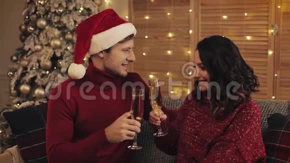 可爱的年轻夫妇坐在沙发上靠近圣诞树在舒适的家背景下闪烁着香槟妻子的眼镜视频的预览图