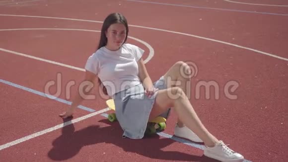 漂亮的女青年穿着短裙坐在滑板上看着篮球场上的摄像机视频的预览图