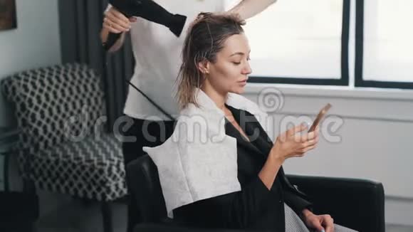 关闭理发师用吹风机擦干女性湿发客户使用手机视频的预览图