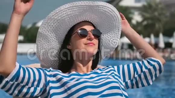 带着太阳镜和白帽子在游泳池附近晒日光浴的轻松微笑的女士中等特写镜头视频的预览图