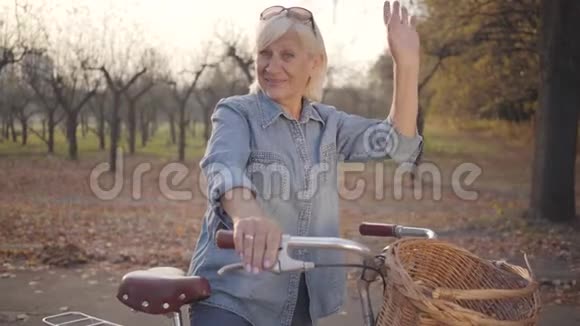 快乐成熟的白种人女士穿着牛仔布站着骑着自行车对着镜头挥手美丽的欧洲退休妇女视频的预览图