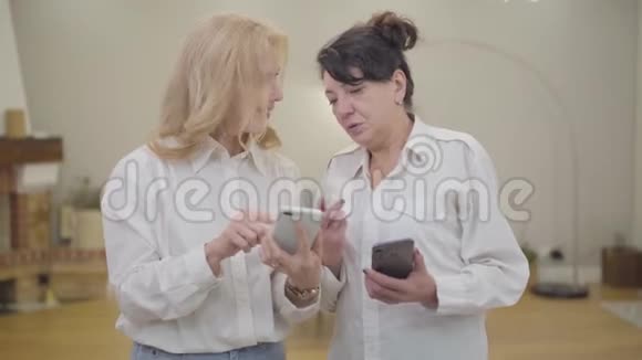 成熟的白人女性用智能手机教她的朋友两位穿白色衬衫的高级女士的肖像视频的预览图