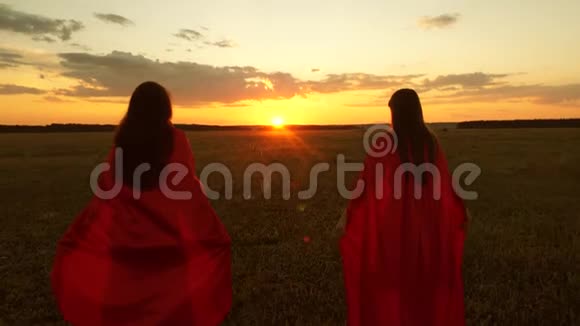 穿红色斗篷的女孩在日落时扮演超级英雄团队合作女朋友在夕阳下跑过傍晚的田野视频的预览图