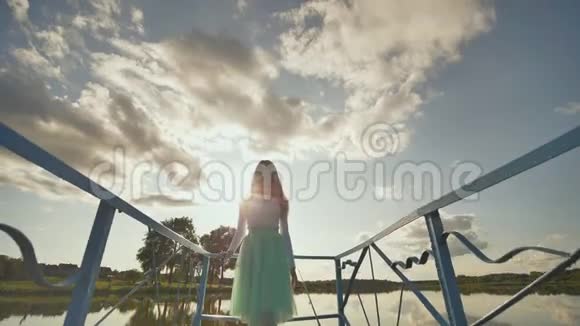 日落时一个穿着蓝色裙子的快乐女孩沿着湖上的桥跑来跑去录像在移动视频的预览图