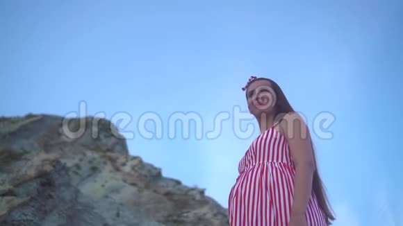 年轻的怀孕女孩穿着条纹红白连衣裙在岩石山脚下显示出自由的迹象举手视频的预览图