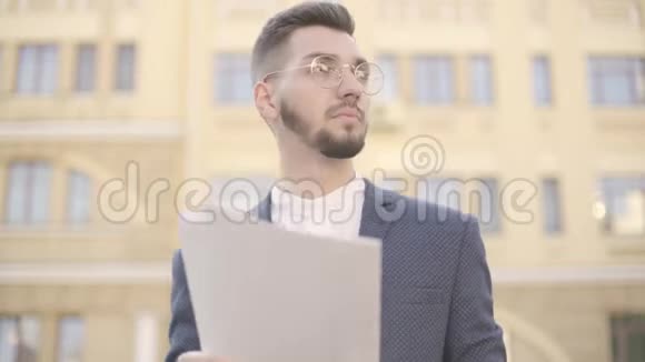 戴眼镜的年轻穿着考究的商人站在高楼大厦前阅读报纸的肖像办公室视频的预览图