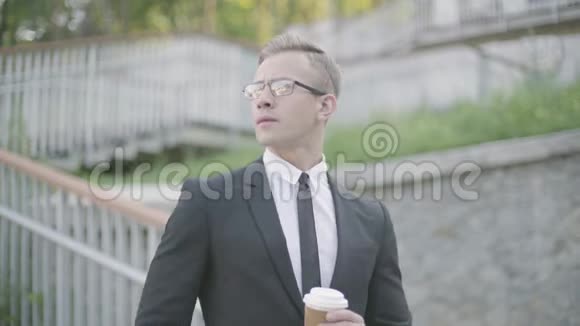 一幅英俊自信的年轻商人戴着眼镜站在楼梯前喝着咖啡看着视频的预览图