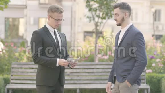 两个年轻的衣着讲究的伙伴在公园里见过面自信的经理数着钱把钱给了那家伙视频的预览图