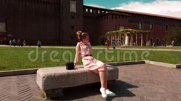 疲惫的白种人女孩坐在石凳上在晴朗的晴天休息在堡垒墙的背景下视频的预览图