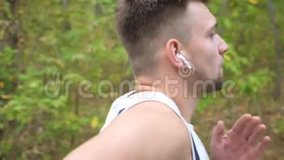 在森林附近的小径上年轻强壮的运动员紧紧地迎着他快速冲刺带无线耳机的运动员视频的预览图