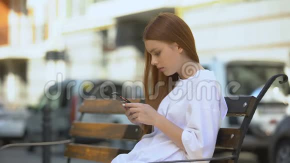 在长凳休息时一个严肃的少女在智能手机上滚动玩电子游戏成瘾视频的预览图