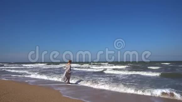 一个身材苗条的女人穿着一件长裙子迎风飘扬站在沙滩上海浪拍打着她的脚视频的预览图