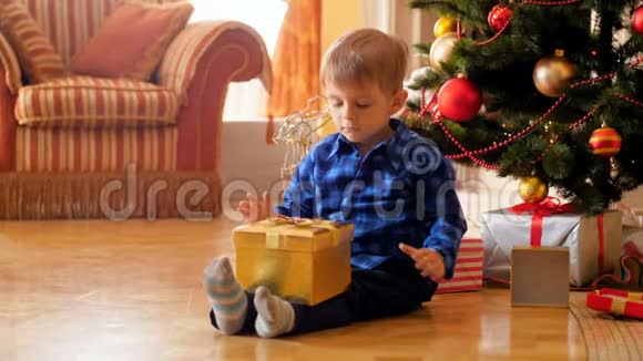 4k视频小可爱男孩坐在圣诞树旁边的地板上打开圣诞老人送礼物的盒子视频的预览图